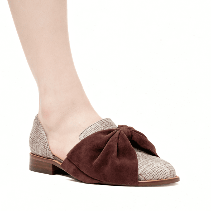 A woman's slip-on shoe with a large dark brown suede bow on the toe, combining a brown suede upper with a beige and gray tweed patterned toe cap and low block heel, shown on a model's foot against a white background.