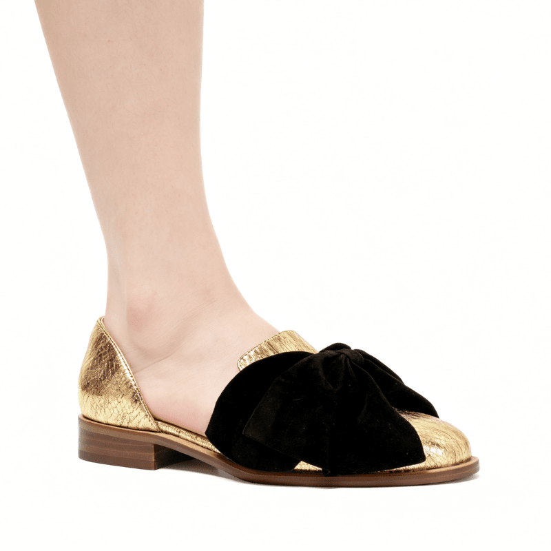 A close-up of a person's foot wearing a flat loafer with a black velvet bow across the toe, a metallic gold toe cap and heel, and a brown sole, shown against a plain white background.