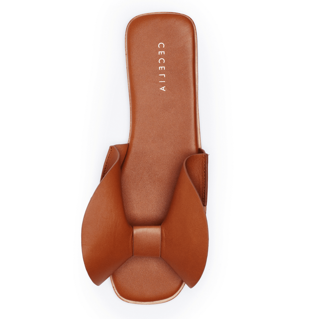 A cognac-colored flat sandal with a voluminous bow on a square toe and cushioned footbed, branded with Cecelia